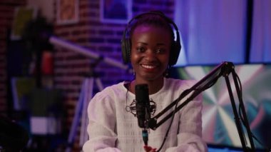 Kayıt stüdyosunda profesyonel ekipman kullanarak canlı yayında kameraya kendinden emin bir şekilde gülümseyen online radyo sunucusunun portresi. Afrika kökenli Amerikalı podcaster mikrofonuyla masada oturuyor..
