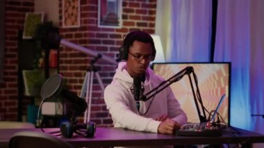 Afrikalı Amerikan Podcaster 'ın portresi mikrofon kullanarak dinleyicilerle konuşurken dijital ses mikserini ayarlıyor. Erkek online radyo sunucusu ev stüdyosunda kayıt yaparken kameraya kendinden emin gülümsüyor..
