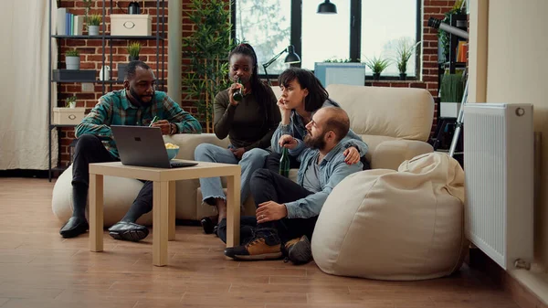 Multiethnic group of people having conversation and drinking beer, enjoying leisure activity at home. Friends bonding over memores, talking about friendship and laughing together.