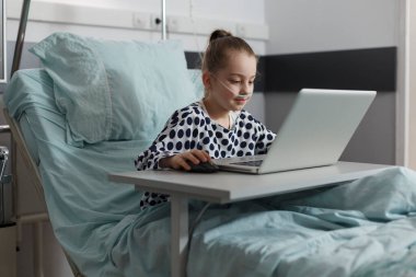 Pediyatri kliniğinin çocuk koğuşunda yatarken laptopta oyun oynayan hasta küçük kız. Modern bilgisayarda oyun oynarken hasta yatağında dinlenen hasta bir çocuk..