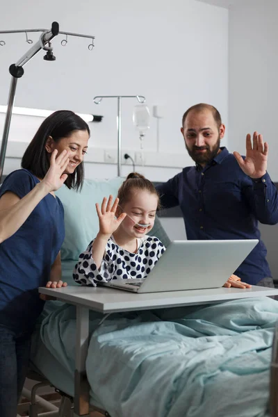 Sick little girl in healthcare facility talking with relatives on virtual online call on modern computer. Caring parents sitting beside ill kid while waving at video call on laptop.