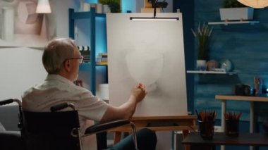 Fiziksel engelli bir ihtiyar tuvalde sanat ve zanaat hobisi için resim çiziyor. Tekerlekli sandalye kullanıcısı sanatsal tasarım modelinin profesyonel taslağını oluşturuyor.
