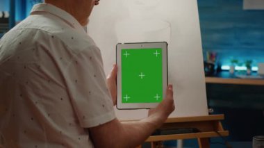 Yaşlı adam yeşil ekran arka planı olan dijital tablet kullanıyor ve sanat aletleriyle başyapıt çiziyor. Boş krom anahtar şablonuna sahip telefon tutuluyor, izole edilmiş kopyalama alanı.