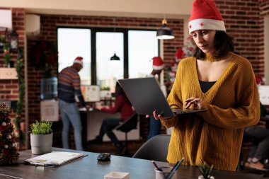 İşkadını ofiste dizüstü bilgisayarını dekorasyonla kullanıyor, Noel arifesini kutlamak için meslektaşlarıyla birlikte bilgisayar başında çalışıyor. Xmas süslemeleri ve iş yerinde ağaç.