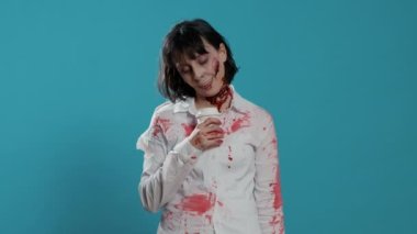 Derin yaraları olan ürkütücü görünümlü zombi mavi arka plandaki bir fincandan kahve kokusu alıyor. Korkunç çılgın görünümlü, kanlı yaraları olan bir canavar kameraya hırıldarken bir fincan kahve içiyor..