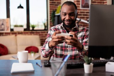 Genç yetişkin cep telefonuyla video oyunu oynuyor, online oyun yarışmalarıyla eğleniyor ve evden çalışıyor. Akıllı telefondan oyun oynamak eğlenceli, serbest çalışan serbest etkinlik yapıyor..