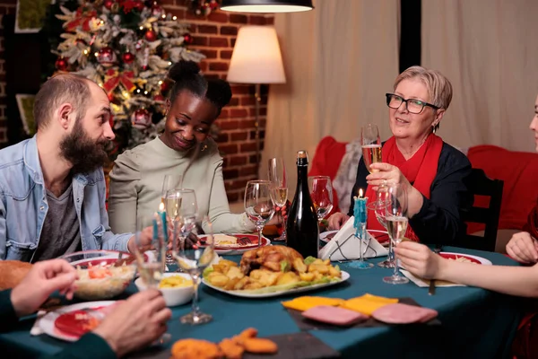 Smiling woman proposing christmas toast at family gathering, holding glass with sparkling wine at festive table. Diverse couple celebrating winter holiday with parents, drinking at new year party