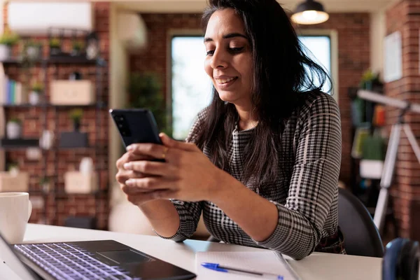 Female freelancer working on smartphone app and using laptop for remote job at home desk. Browsing internet and social media on mobile phone to text messages, online research break.