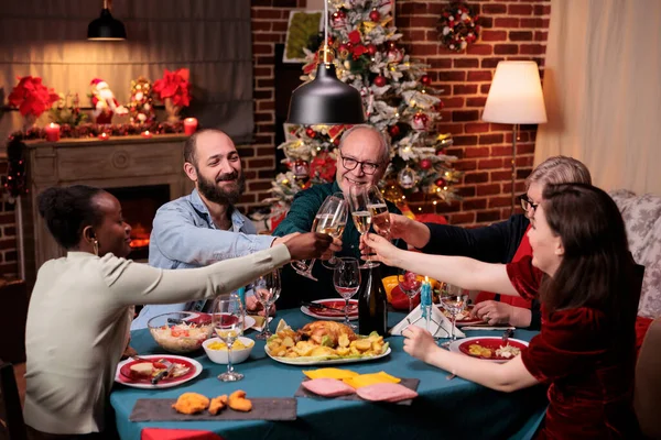 Diverse family clinking glasses with sparkling wine, proposing christmas toast, drinking at festive dinner. Winter holiday celebration with parents, people gathering together
