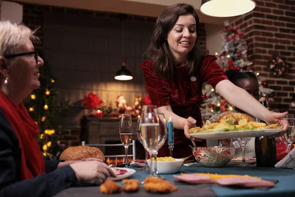 Chrismas family home party, diverse friends gathering at festive dining table, eating traditional dinner together, winter holiday celebration. Woman holding chicken dish, people celebrating xmas