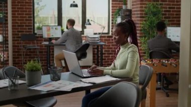 African american woman working on laptop in business office, analyzing charts on presentation to do research and paperwork. Company employee sending email to management department.