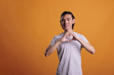 Genç, mutlu Asyalı adam kalp şeklinde aşk jesti, aşk sembolü gösteriyor. Gülümseyen neşeli genç takdir işareti yapıyor, pozitif yüz ifadesiyle kameraya bakıyor.