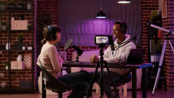 African american online radio host recording vlog using digital camera on tripod and intruducing guest to listeners in home studio. Content creator interviewing famous vlogger while streaming live.
