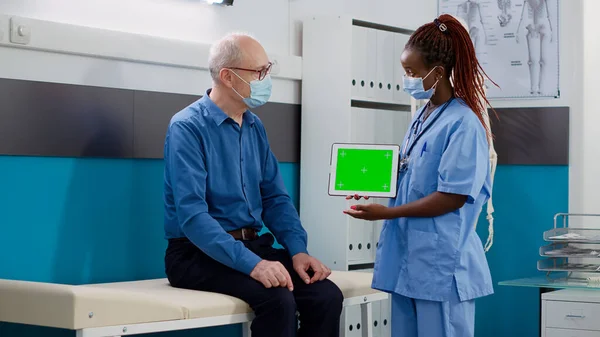 医療キャビネットのシニア男性に緑色の画面でタブレットを示す看護師 パンデミックの間の空白のモックアップテンプレートとコピースペースと隔離されたクロマキーの背景を見る — ストック写真