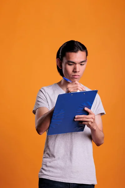 Young asian man filling survey form in clipboard, serious student writing in notepad, taking notes. Focused manager holding papers, secretary making office plan checklist