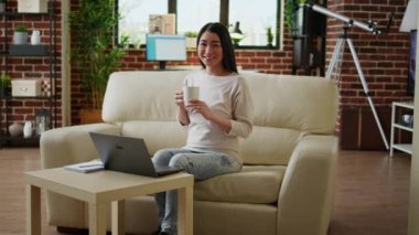 Joyful asian woman sitting in living room working from home while smiling at camera. Happy young adult student inside modern apartment having laptop on table while waiting for online class to start.