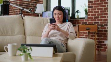 Happy woman browsing social media on smartphone while wearing headphones and working remotely from home. Cheerful smiling heartily person chatting on mobile phone while doing remote work.