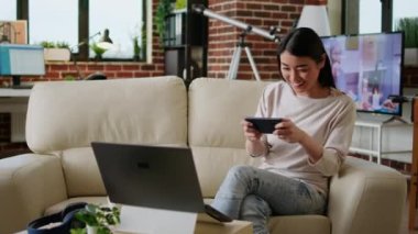 Playful woman sitting on sofa at home while playing arcade games on smartphone. Competitive looking young adult person enjoying mobile gaming while relaxing in living room on couch.