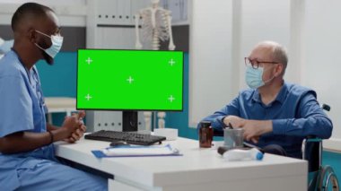 Hemşire ve engelli yaşlı adam kabinedeki yeşil ekrana bakıyor. Tekerlekli sandalye kullanıcısı ve yüz maskeli uzman izole edilmiş krom anahtar görüntüsü ve kopyalanmış kopya alanı kullanıyor.