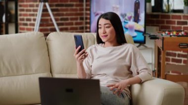 Woman working remotely while in digital videoconference on smartphone with manager. Young adult asian person waving at phone camera while doing remote work and attending videocall with colleague.