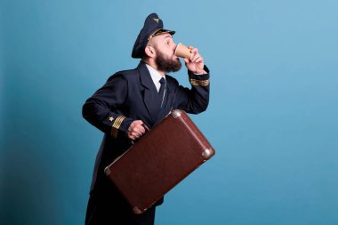 Profesyonel havayolu üniformalı bir kaptan geç kalıyor, bavul taşıyor, paket kahve içiyor. Havaalanında bavulu olan bir pilot, elinde çay fincanı olan bir pilot, yan görüş.