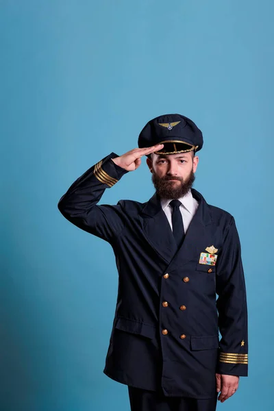 Confident airplane aviator saluting, wearing uniform and hat front view portrait, plane captain looking at camera. Civil captain with aircraft wings badge on jacket, professional aircrew member