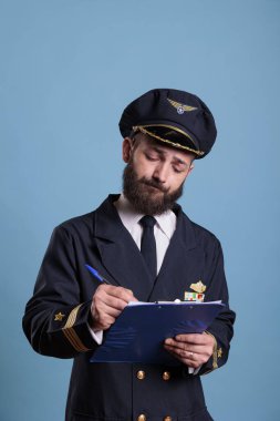 Uçak pilotu pano tutuyor, doldurma formu dolduruyor, belgelerle dolu bir havacı. Üniformalı bir uçak kaptanı, hava alanında ön tarafta yazıyor. Stüdyo orta derece mavi arka planda.