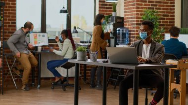 Yüz maskeli bir proje yöneticisi dizüstü bilgisayardaki verilerle çalışıyor, grafikler ve grafiklerle iş sunumu planlıyor. Araştırma bilgileriyle şirket raporu oluşturuluyor. Üçayak atışı.