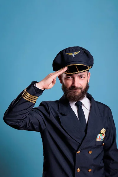 Smiling plane pilot saluting, wearing uniform and hat front view portrait, airplane captain looking at camera. Civil aviator with badge on jacket, aircrew member on blue background