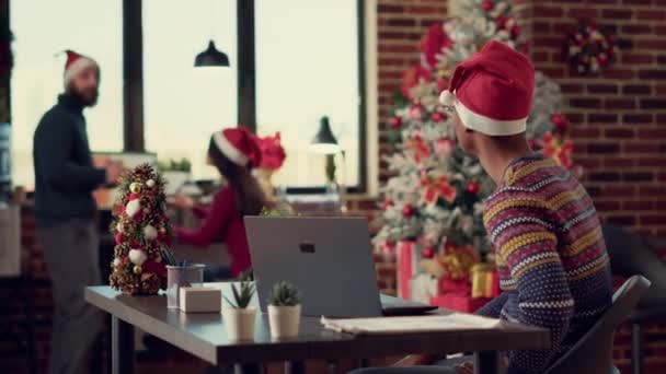 クリスマスの前夜祭の間にコンピュータに関するレポートを分析する人々の多民族チームは サンタの帽子をかぶっている クリスマスツリーや季節の装飾品とオフィスで一緒に働く多様な男性 — ストック動画