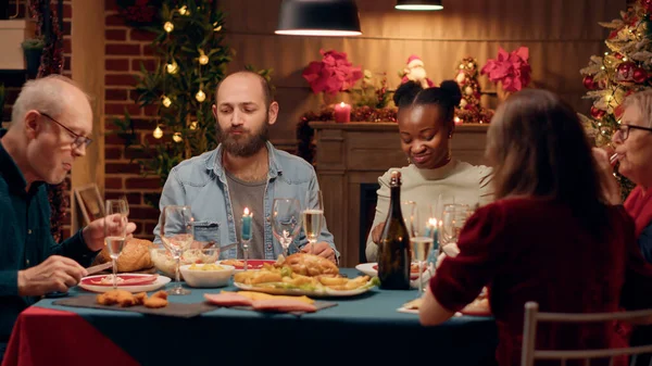 Cheerful multicultural people enjoying Christmas dinner while eating roasted chicken. Happy diverse family members enjoying traditional home cooked food wihle celebrating winter holiday together.