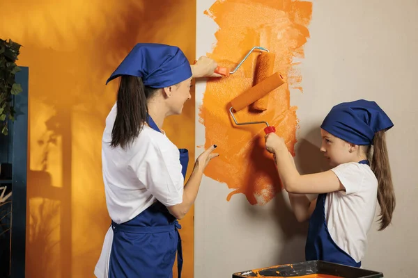 Young people painting walls with orange paint to redecorate apartment space with tools and paintbrush. Woman with little girl renovating house room with roller brush and color, teamwork improvement.