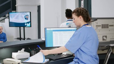 Tesis lobisindeki kayıt masasındaki randevular üzerinde çalışan bir tıp asistanı. Kadın hemşire hastane resepsiyonunda sağlık raporlarını ve formları kontrol ediyor. El kamerasıyla..