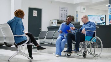 Senior patient in wheelchair doing consultation with nurse in waiting area lobby, examining man with chronic impairment and disability. Specialist and person at checkup visit. Tripod shot.