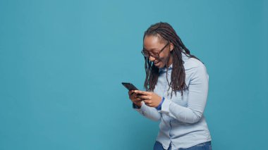 Akıllı telefondaki online uygulamayı kullanarak, sosyal medya internet sitesine göz atarak ve mesajlar hakkında olumlu düşünerek mutlu bir model. Gülümseyen neşeli kişi stüdyoda heyecan gösteriyor..