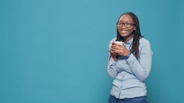 Pozitif bir insan mavi arka planda bir fincan kahve içiyor ve elinde kameranın önünde sıcak bir içecek tutuyor. Mola vermek ve stüdyoda eğlenmek için içki içen mutlu bir kadın..