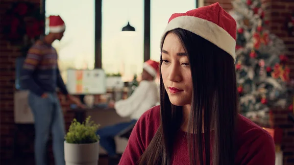 Asian employee working on laptop in decorated office, celebrating christmas holiday at startup job. Festive woman with santa hat using computer at desk, celebrating winter season.