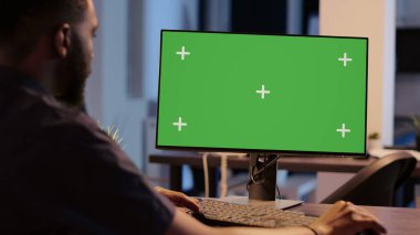 African american man looking at computer with greenscreen, analyzing isolated display on desktop monitor. Using blank copyspace template with mockup screen and chroma key background.