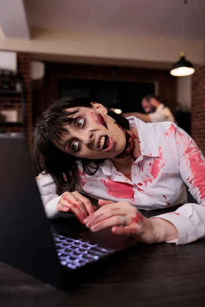 Scary horrific zombie using laptop at desk, undead corpse trying to work on computer in startup office. Creepy aggressive brain eating monster looking terrifying and horrible, sinister danger.