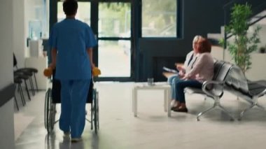 Hemşire tekerlekli sandalyedeki engelli hastaya destek veriyor, tıbbi danışmanlık muayenesine katılıyor. Sağlık kliniğinde kronik ve fiziksel engelle uğraşan Asyalı bir kadın. El kamerasıyla..