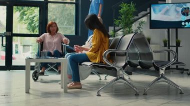 Engelli bir Asyalı kadın hastane lobisinde yaşlı bir doktorla konuşuyor, tekerlekli sandalye kullanıcısıyla tıbbi danışmanlık yapıyor. Klinikte kronik fiziksel engelli bir hasta. Üçayak atışı.