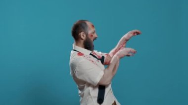 Scary looking brain-eating doing a funny dance on blue background. Spooky brain dead zombie with deep aned bloody wounds and scars dancing goofy while looking at camera. Studio shot