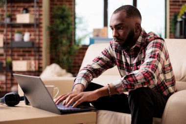 Afrikalı Amerikalı bir adam interneti dizüstü bilgisayarla tarıyor, sosyal medya uygulaması üzerinde çalışıyor ve internet üzerinden iş raporu hazırlıyor. Bilgisayarda uzaktan bağımsız çalışmak ve yönetim sunumunu analiz etmek.