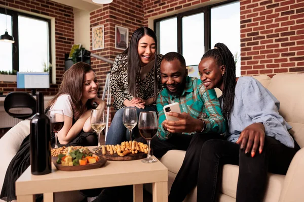 在客厅的生日派对上 多种族人群在智能手机屏幕上看有趣的视频 吃开胃菜 面包棒 — 图库照片
