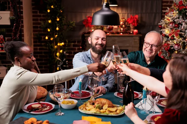 Family proposing christmas toast at festive dinner, clinking glasses with sparkling wine at xmas home feast. Winter holiday celebration with parents, drinking at new year party