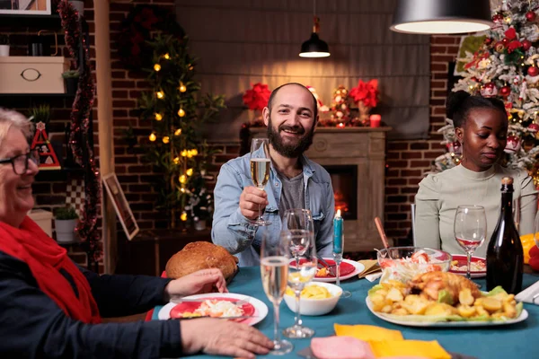 Family christmas celebration, friends drinking at festive served dinner table. Smiling man holding sparkling wine glass, happy diverse people eating xmas dishes, winter holiday home evening party