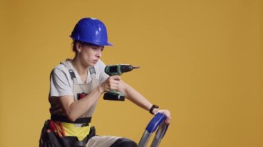 Kendine güvenen inşaatçı çivi çakmak için elektrikli matkap kullanıyor. İnşaat yenileme için mühendislik makinesiyle çalışıyor. Sarı arka planda çivi tabancasıyla sondaj yapan bir kadın..