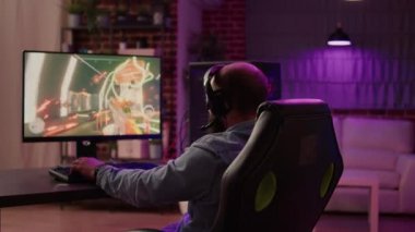 Over shoulder view of man playing online action game congratulates team on headset after celebrating victory on gaming pc at home. Professional gamer winning tournament doing victory hand gesture.