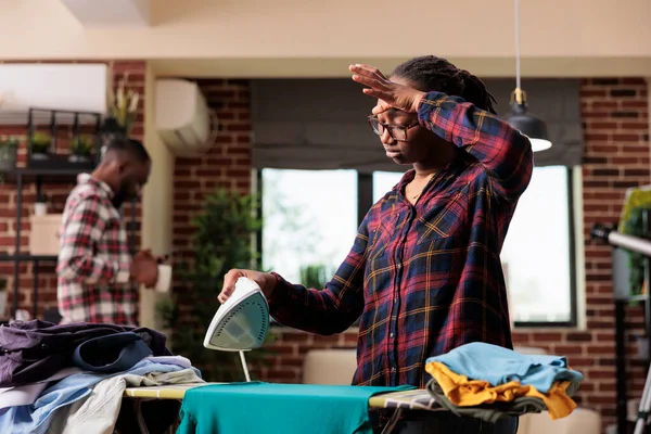 Nieszczęśliwa Afroamerykanka Prasująca Ubrania Podczas Gdy Zrelaksowany Mąż Używa Telefonu — Zdjęcie stockowe
