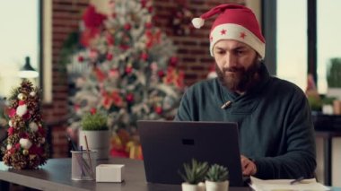 Genç yetişkin planlama raporu kış tatili sezonunda dizüstü bilgisayarda festival ofisinde bilgisayar üzerinde çalışıyor. Noel Baba şapkalı bir adam Noel arifesinden önce araştırma yapıyor. Üçayak atışı.